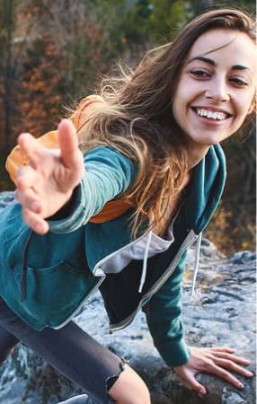 Woman hiking on a rocky trail, smiling and reaching out a hand to help someone climb up, with autumn trees in the background.