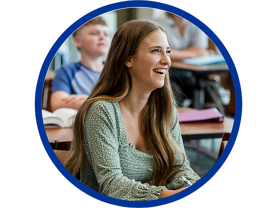 Smiling teenage girl with long hair sitting at a desk in a classroom, engaged and listening, with classmates and open books in the background