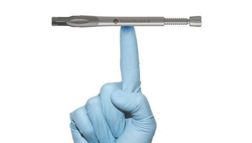 Hand wearing a disposable glove with the index finger pointing upwards. The Clareon Monarch IV Handpiece sits horizontally, balanced on top of the index finger.
