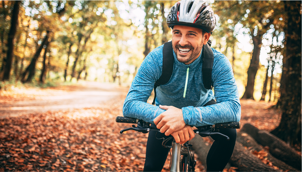 A smiling man outside in a blue hoodie sitting on his bike and leaning forward on the bike handles. The man has a black backpack and is wearing a black and gray helmet. Colorful leaves appear on the ground around him indicating that it is fall.