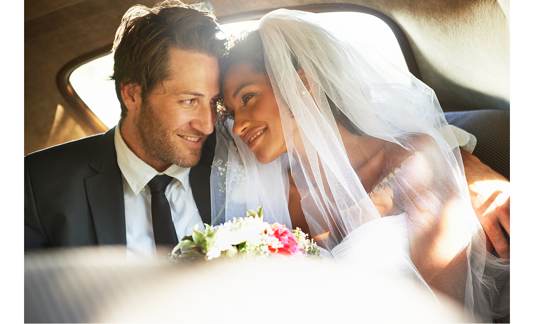 A married couple sitting side by side in a car. They are touching foreheads while smiling at each other. The man is wearing a black suit, with a white shirt and black tie. The woman is wearing a white wedding dress. A bouquet of white and pink flowers appears between them.