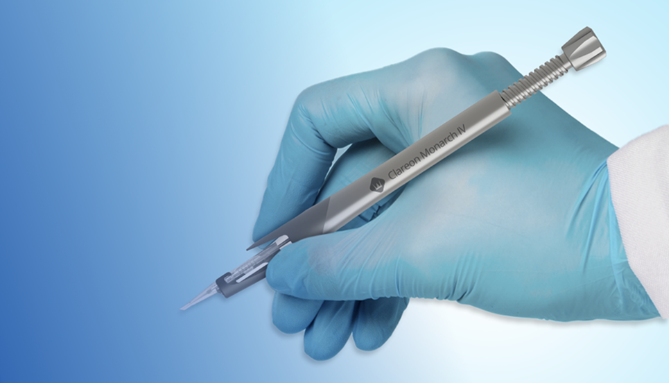 Een afbeelding van de Clareon Monarch IV Delivery System, vastgehouden door een hand in een blauwe chirurgische handschoen. De hand houdt het apparaat vast zoals men een pen zou vasthouden en verschijnt tegen een blauwe achtergrond.
