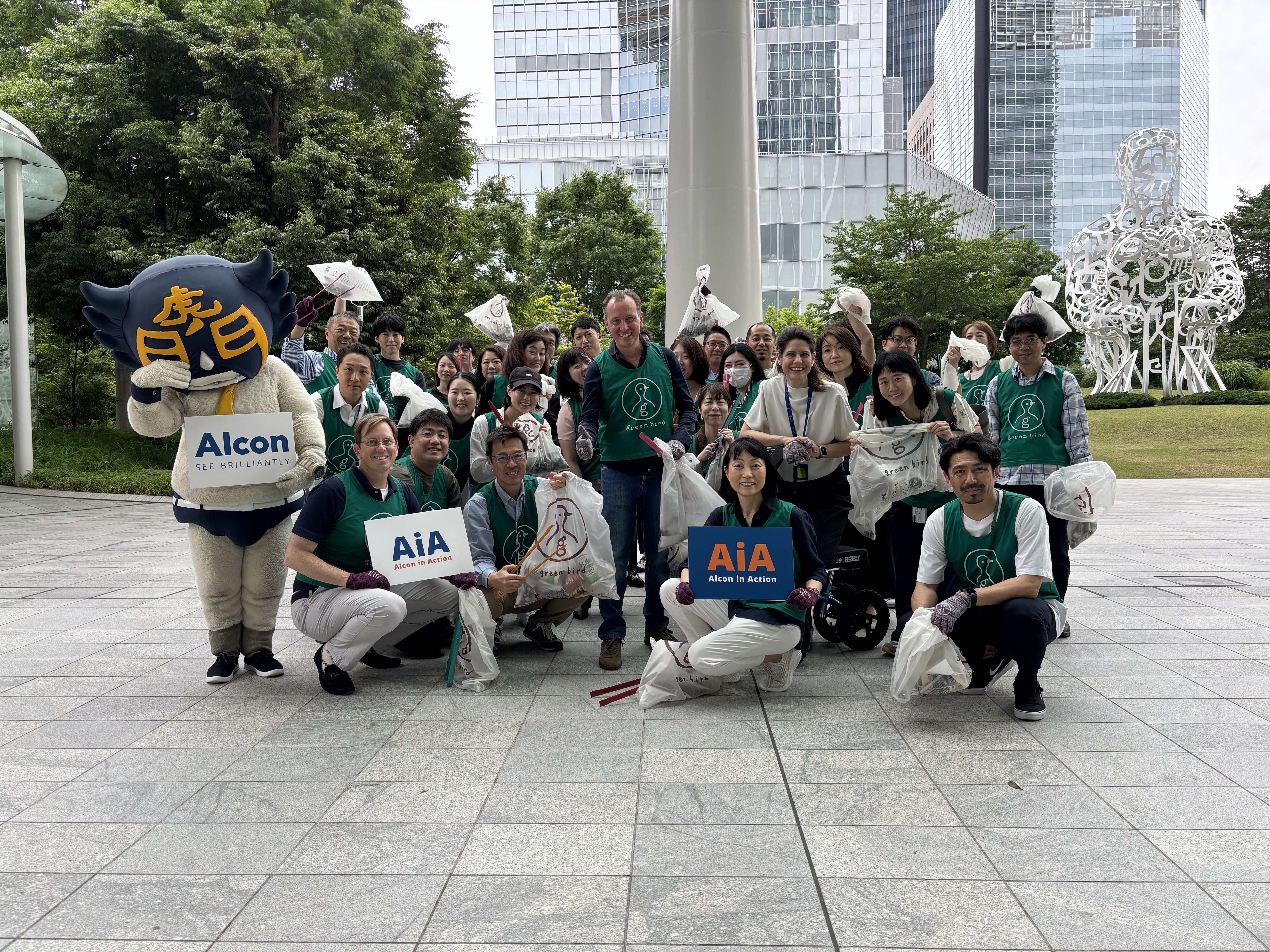 「Alcon」および「AiA – Alcon in Action」の看板を持ち、ゴミ袋を手にしたアルコンの社員たちが屋外で集合写真を撮影。参加者は緑色のベストを着用し、左側にはマスコットキャラクターが立っている。都市公園での清掃活動の様子。
