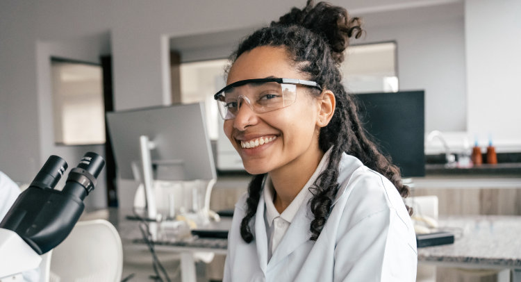 A young Black woman wearing safety glasses and a lab coat is smiling at the camera. She is in a laboratory setting with scientific equipment visible in the background.