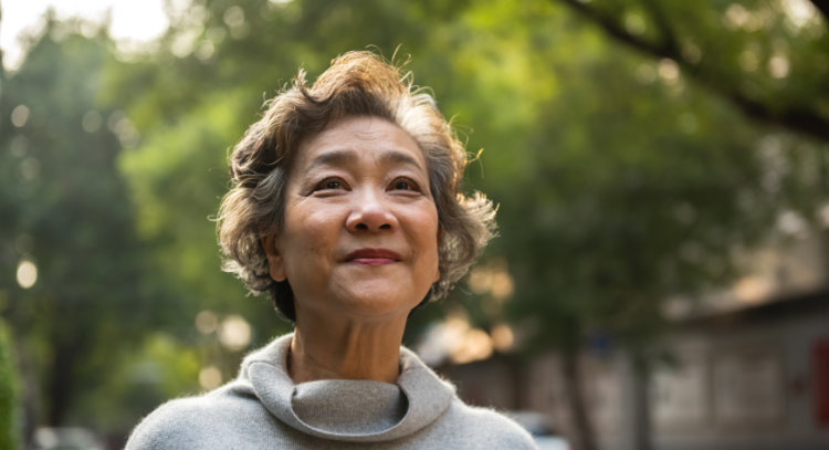 A mature Asian woman with short, graying hair is smiling and looking up towards the trees. She is wearing a gray sweater.