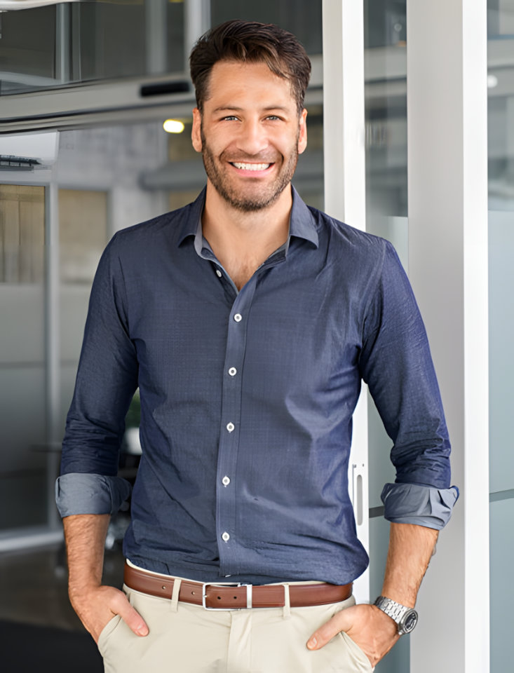 A smiling man with short brown hair is leaning against a glass wall in a modern office space. He is wearing a blue shirt and khaki pants.