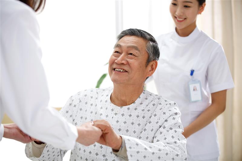 Smiling man in hospital gown grasping hands with doctor with nurse smiling behind him