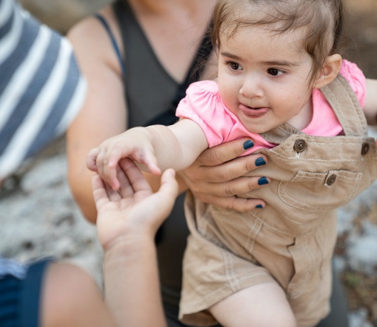  Dospelý s modrými nechtami drží dieťa s tmavými vlasmi, oblečené v ružovom a hnedom. V pozadí je textúrovaný povrch. 