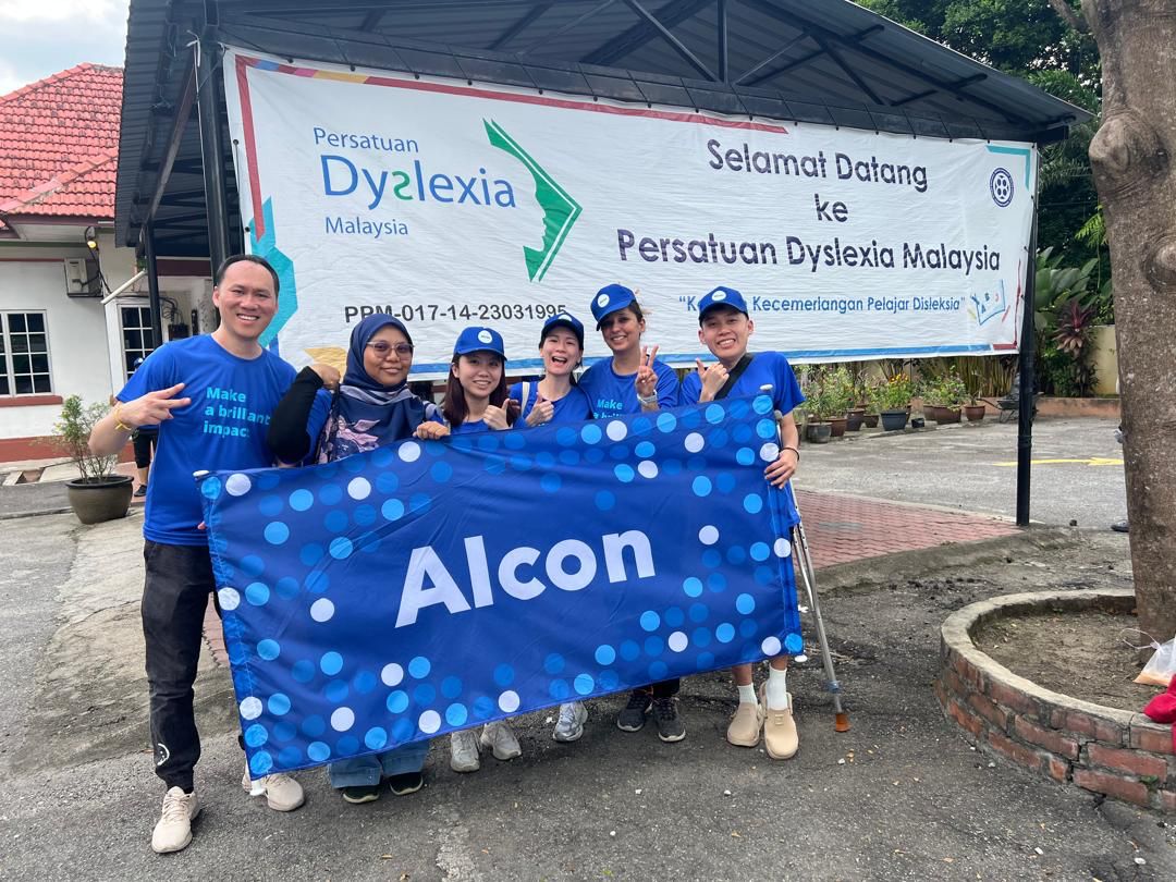 Group of Alcon employees wearing Alcon blue shirt, hats and head coverings, standing side-by-side and holding an Alcon banner