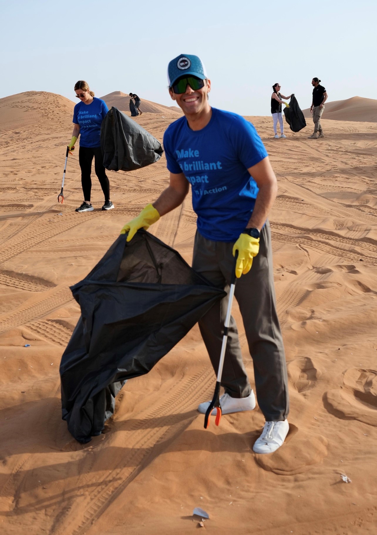  Alcon employees in blue shirts, with trash bags and grabber devices, cleaning up trash in the sand