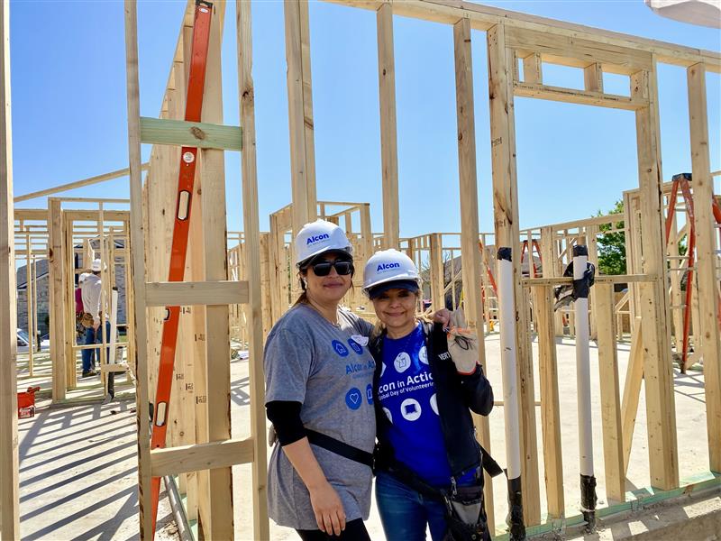 Two Alcon employees wearing Alcon in Action t-shirts, standing in front of the framework for a home being built by Habitat for Humanity