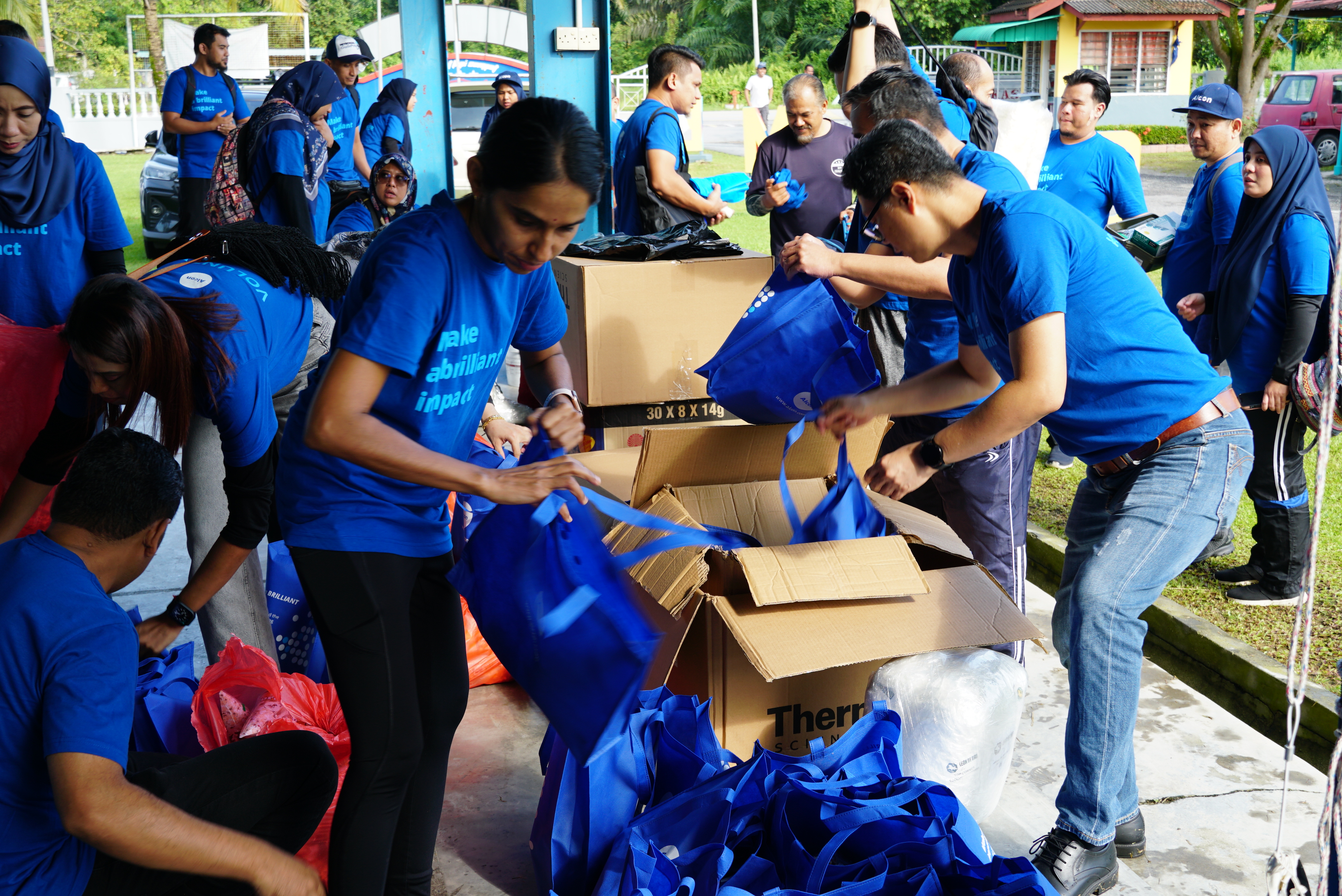 Group of Alcon employees in Alcon blue t-shirts, hats and head scarves, filling blue reusable bags with items to give to people in need.