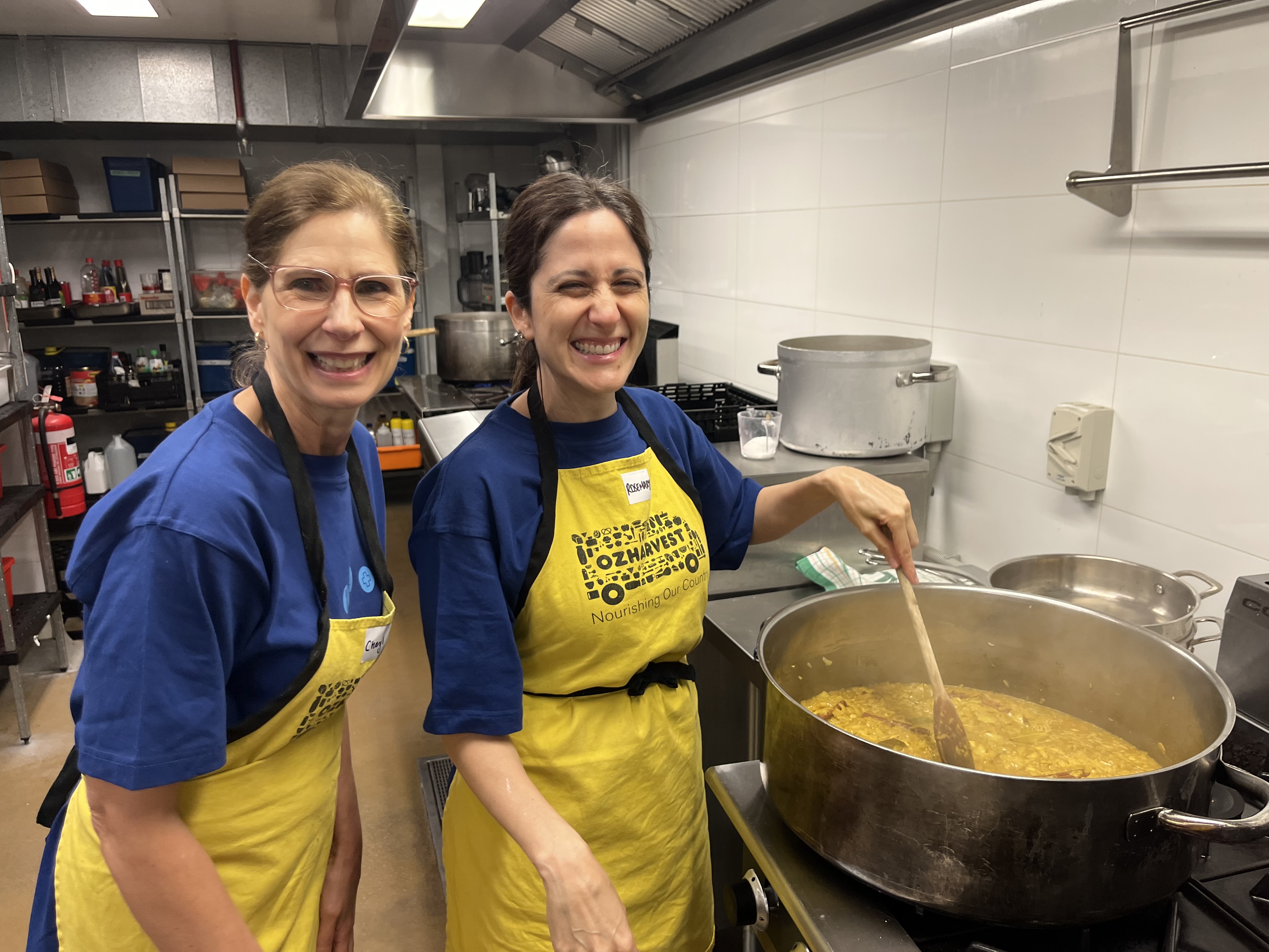  Two Alcon employees in a professional kitchen, wearing yellow aprons and smiling for the camera. One is stirring a large pot of food on a stove.