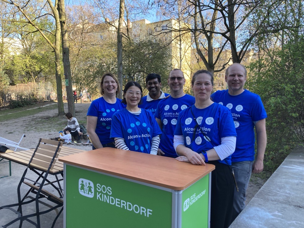 Group of Alcon employees in Alcon in Action blue t-shirts, standing behind behind a booth with "SOS Kinderdorf" on the front