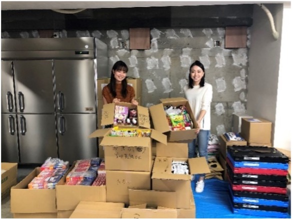 お菓子や物資の詰まった段ボール箱を整理・仕分けしながら笑顔で写る2人の女性。寄付や地域支援活動の一環とみられる。背景には業務用冷蔵庫とコンクリート壁がある。