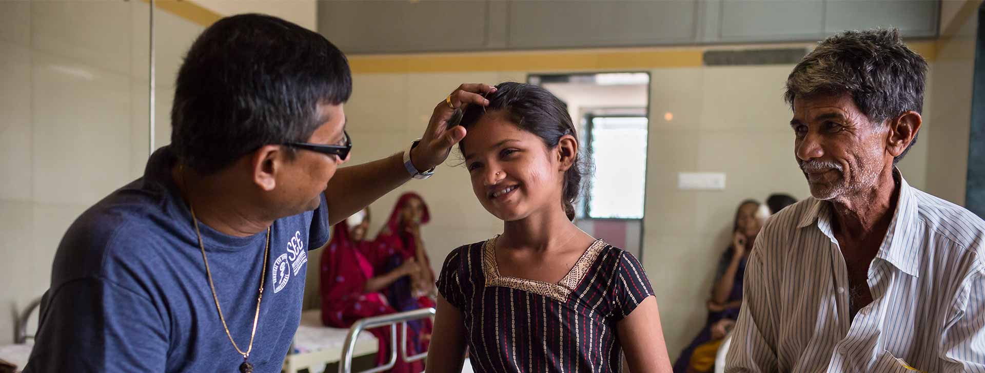 病院または診療所で、笑顔の男性が少女の頭に優しく触れている様子。少女も笑顔で応えている。背景には伝統衣装を着た女性たちが座っている。
