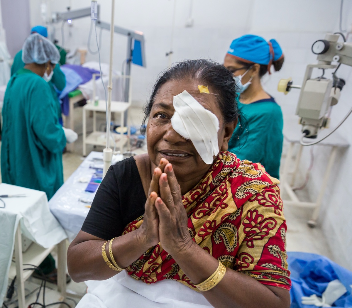 A smiling woman wearing a traditional red and gold sari and gold bangles has an eye patch covering her right eye. She is standing in an operating room, with medical staff in scrubs and surgical caps visible in the background.