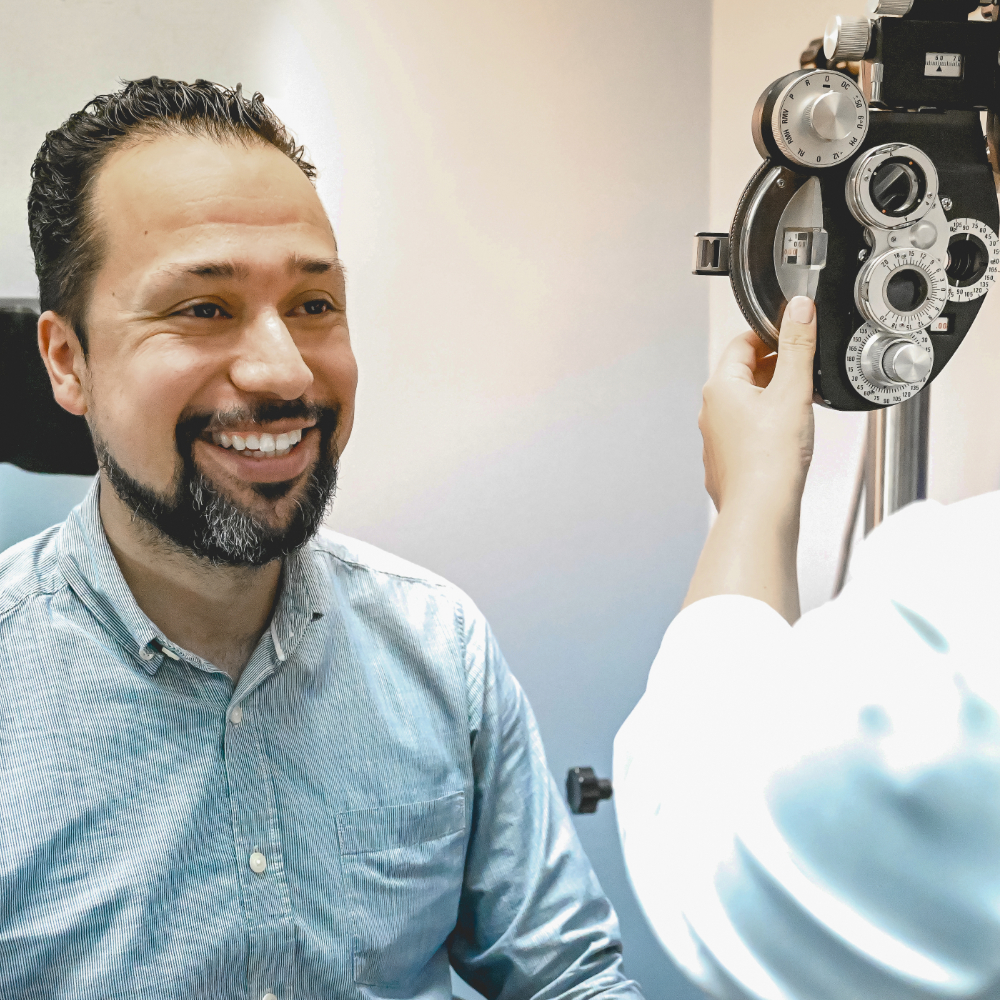 A man is smiling while an optometrist performs an eye exam using a phoropter machine.