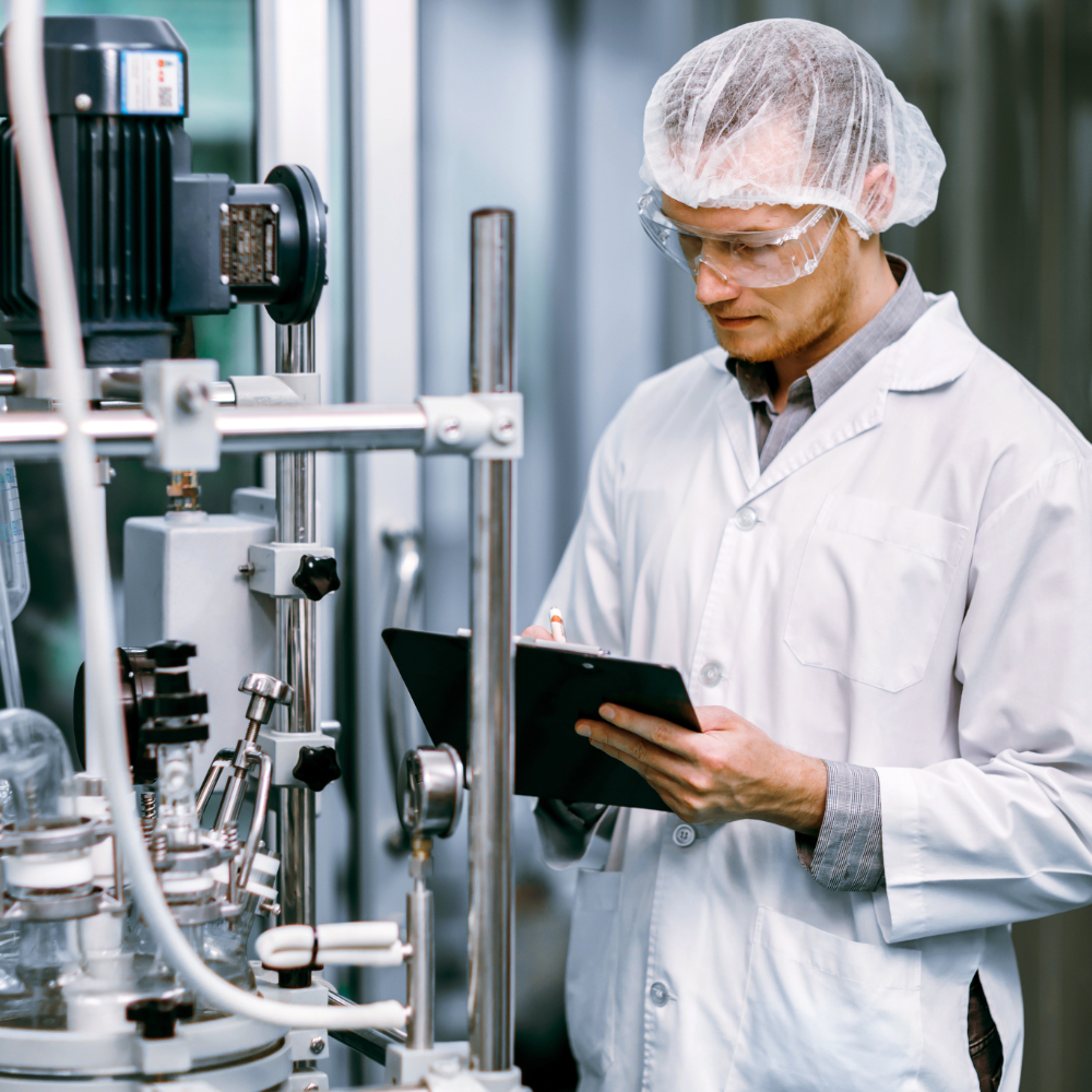 A scientist in a lab coat and hairnet is looking at a tablet while standing next to complex industrial machinery, likely monitoring a process or reviewing data.