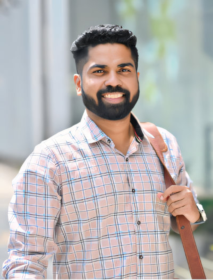A man with a dark beard and short dark hair is smiling at the camera. He is wearing a light-colored plaid shirt and has a brown strap or bag slung over his shoulder.