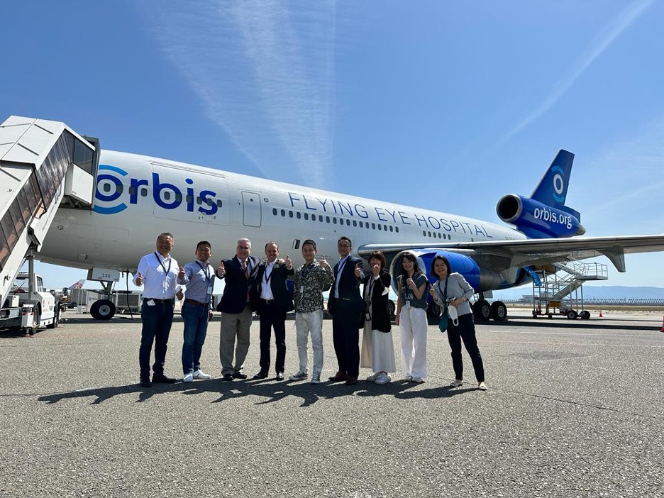 晴れた日に空港の滑走路で、オービスのフライング・アイ・ホスピタルの航空機の前に立ち、笑顔で写真に写る人々のグループ。