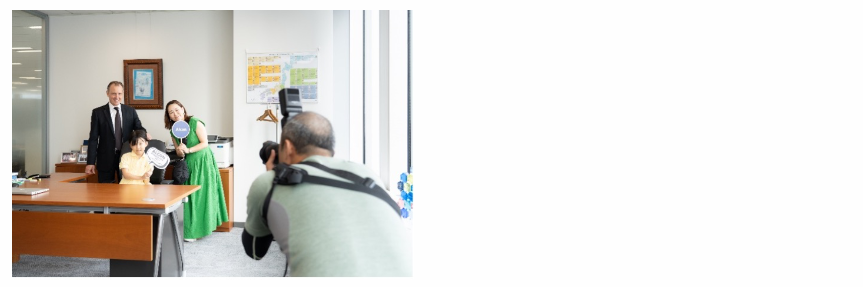 オフィスのような場所で、スーツ姿の男性、黄色いドレスの子供、そして緑のドレスを着た女性が、カメラを持った男性に写真を撮られています。