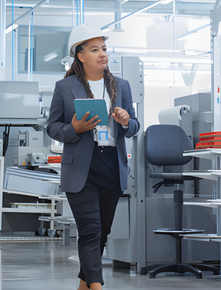 A woman wearing a hard hat, blazer, and dark pants walks through a modern factory. She is holding a small notebook and appears to be inspecting the equipment.