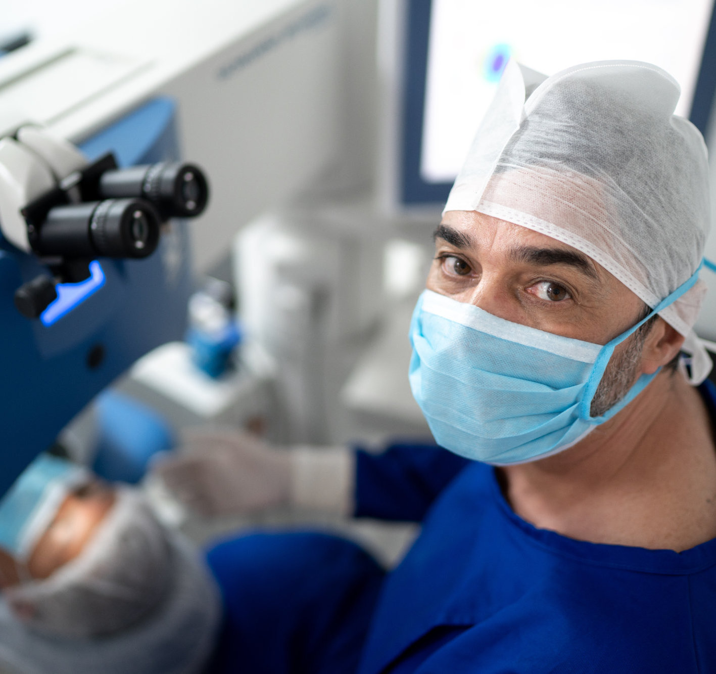 A close-up of an eye surgeon wearing a surgical cap and mask, looking directly at the camera with an eye scope in the background.