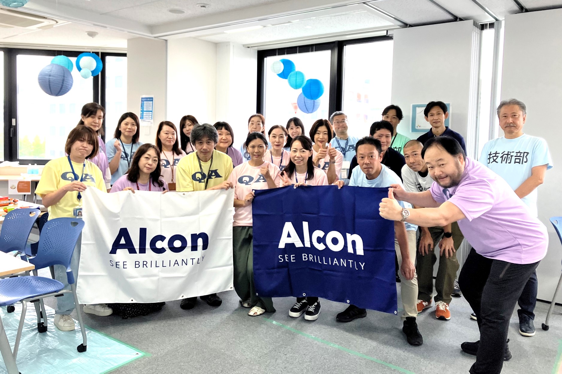 「See Brilliantly」と書かれたアルコンのバナーを持ち、オフィス内で笑顔で集合写真を撮るアルコンの社員たち。背景には青い装飾が見られます。