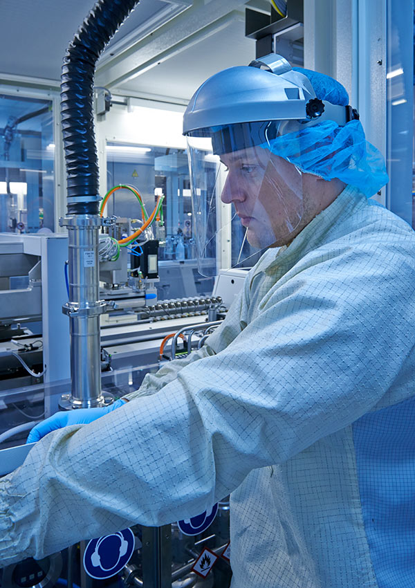 Techniker in vollständiger Schutzausrüstung, einschließlich Gesichtsschutz und Haarnetz, bedient spezialisierte Maschinen in einer Reinraumumgebung.