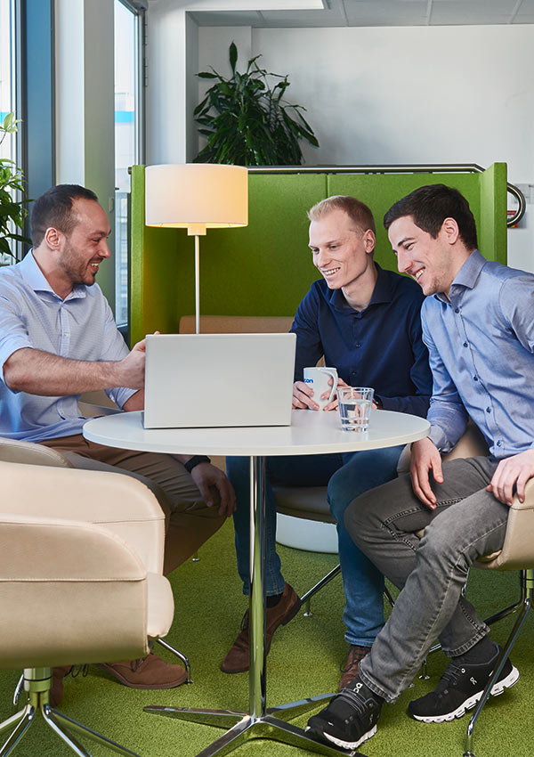 Drei Kollegen sitzen an einem Tisch in einer modernen Bürolounge, lächeln und arbeiten zusammen, während sie auf einen Laptop schauen.