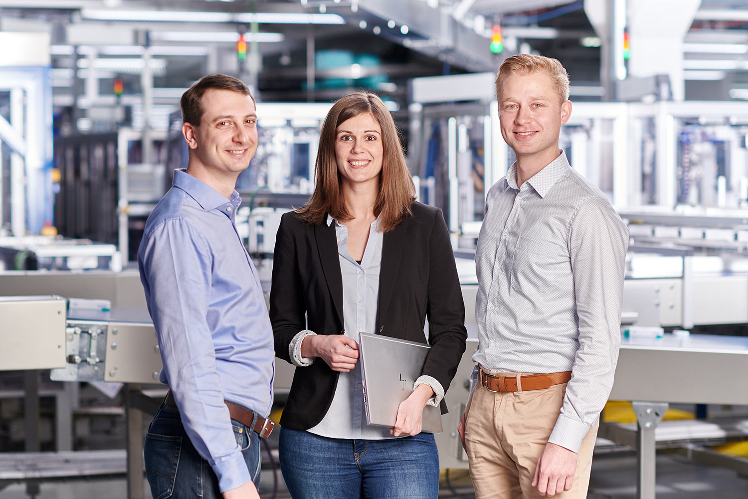 Drei Kollegen stehen zusammen und lächeln in einer modernen Produktionsanlage.