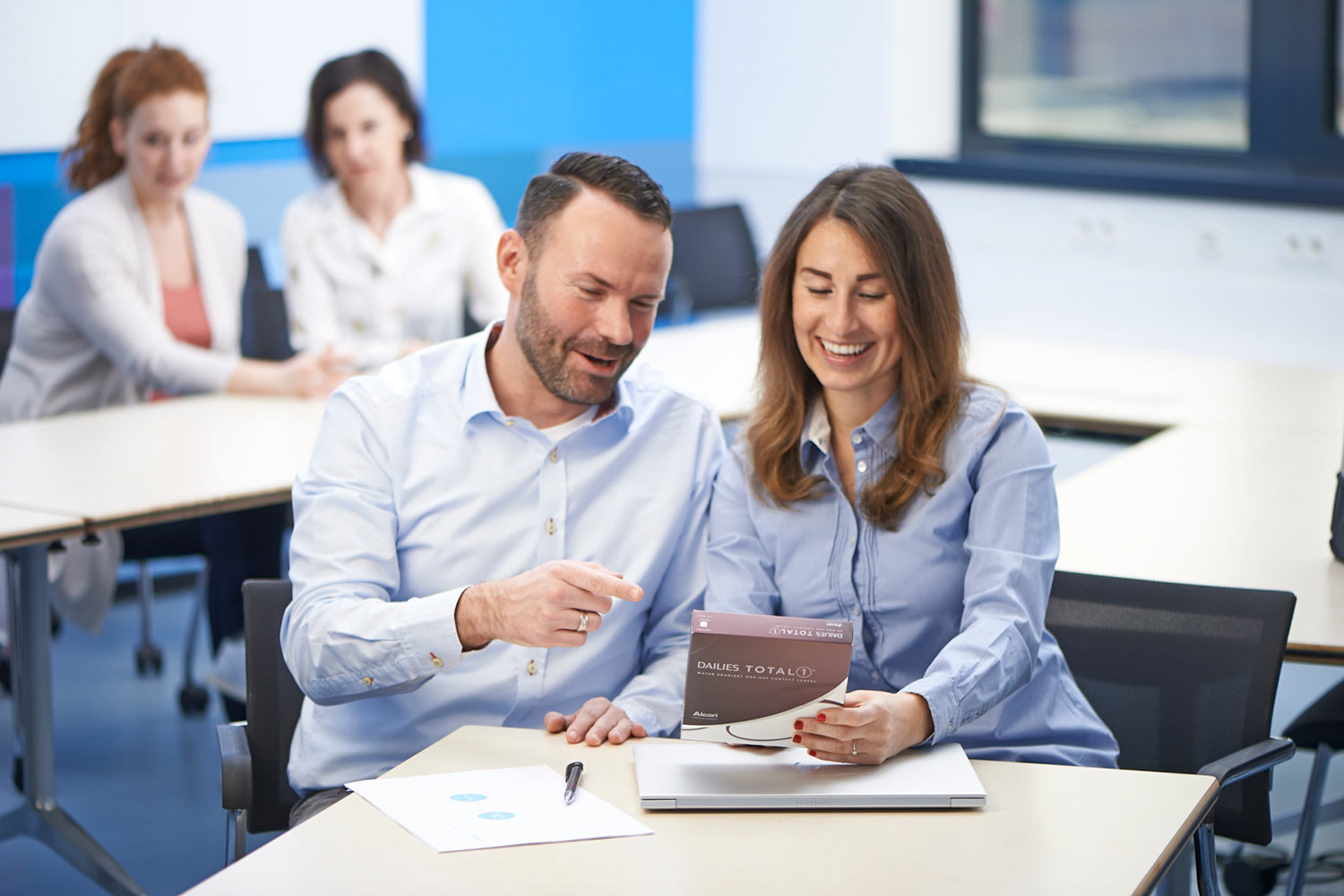 Zwei Kollegen lächeln, während sie gemeinsam eine DAILIES TOTAL1 Kontaktlinsenverpackung in einem Büro betrachten.