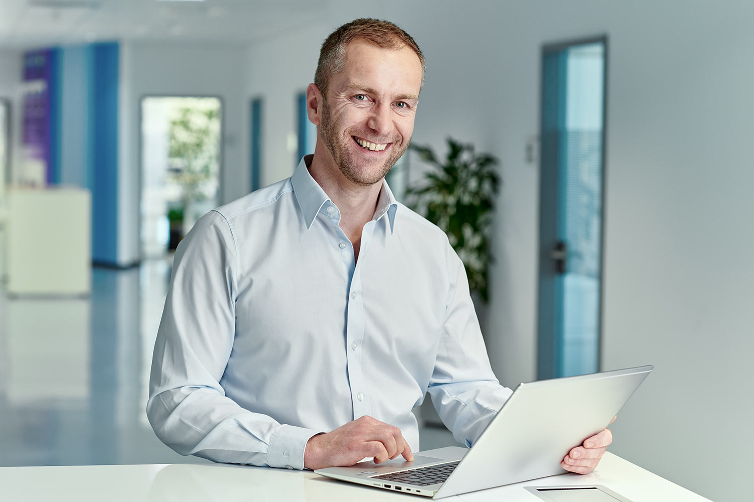 Lächelnder Mitarbeiter im hellblauen Hemd arbeitet an einem Laptop in einem modernen Büroflur.