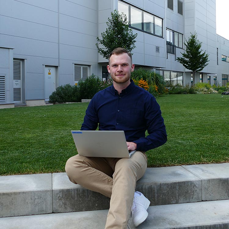Mann sitzt draußen auf Steinstufen mit einem Laptop auf dem Schoß, vor einem modernen Bürogebäude und angelegtem Garten.
