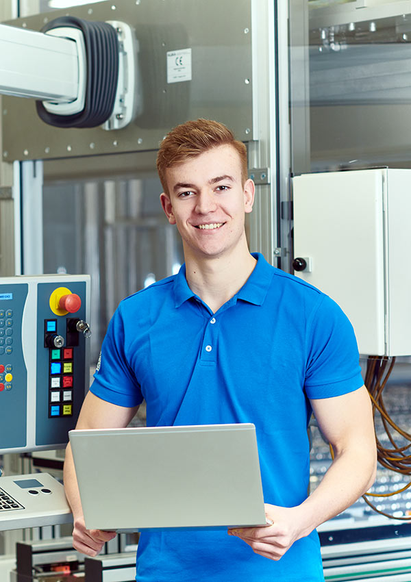 Junger Mann in einem hellblauen Polohemd steht in einer Fabrikumgebung, lächelt und hält einen Laptop. Im Hintergrund sind Steuerpulte und Maschinen zu sehen.