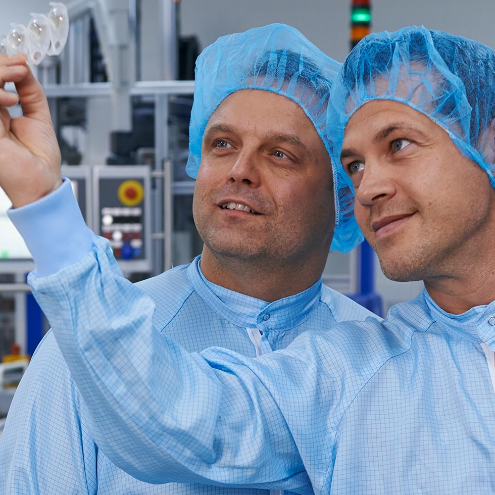 Zwei Männer in blauer Schutzkleidung und Haarnetzen untersuchen in einer Laborumgebung ein durchsichtiges Objekt. Sie wirken konzentriert und engagiert und vermitteln Teamgeist.