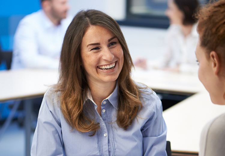 Lächelnde Frau im blauen Hemd im freundlichen Gespräch während eines Meetings in einer hellen Büroumgebung.