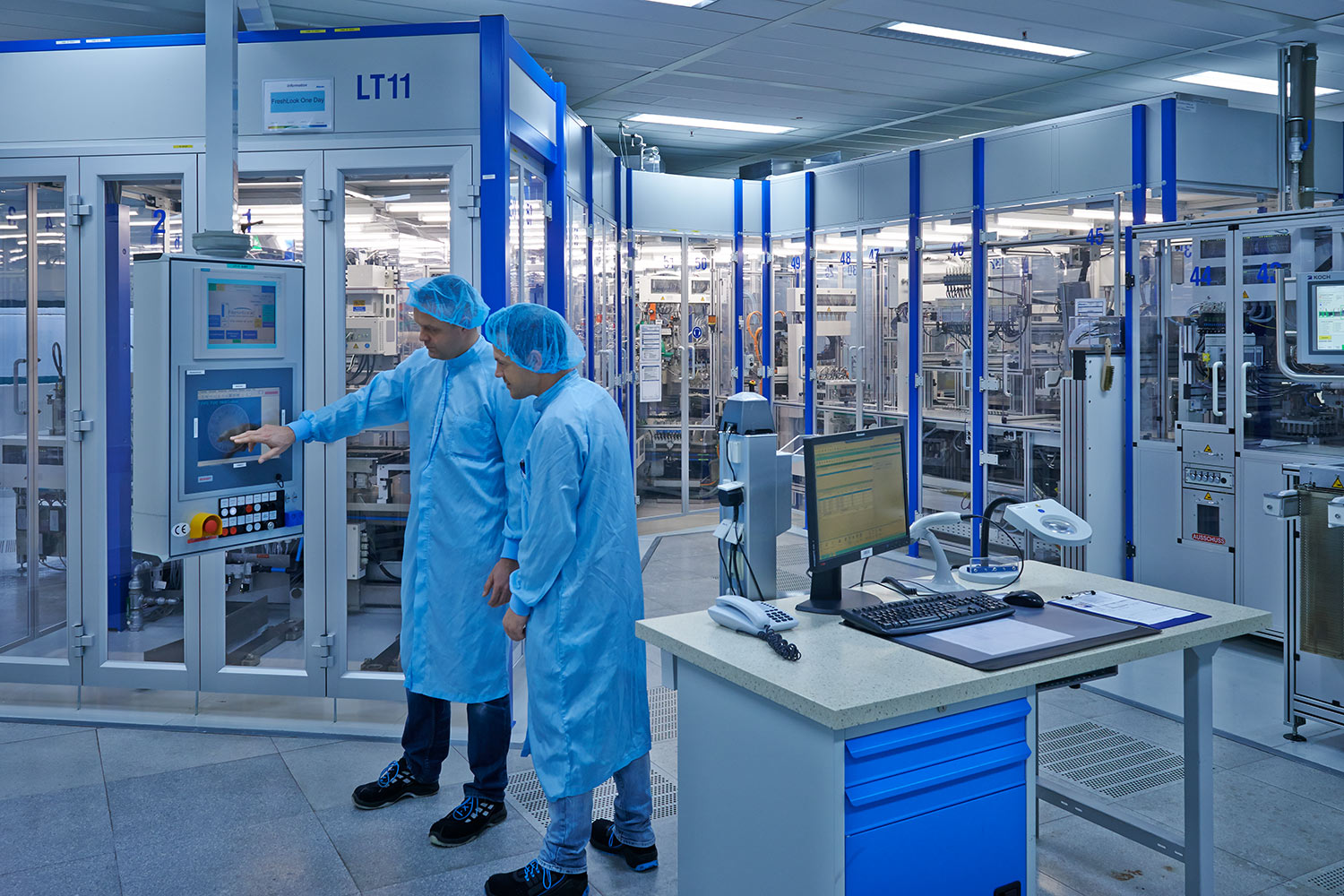 Zwei Mitarbeiter in Schutzkleidung und Haarnetzen bedienen Maschinen in einem Reinraum einer Hightech-Produktionsanlage.