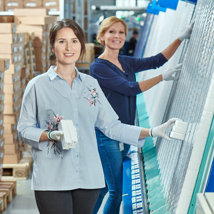 “Zwei Frauen arbeiten in einer Alcon-Produktionsstätte, tragen Handschuhe und sortieren Bauteile an einer großen, geordneten Lagerwand.”