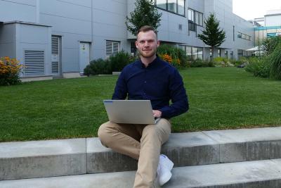 Mann sitzt draußen auf Steinstufen mit einem Laptop auf dem Schoß, vor einem modernen Bürogebäude und angelegtem Garten.