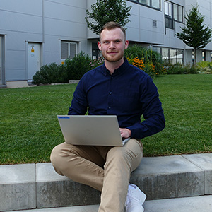 Mann sitzt draußen auf Steinstufen mit einem Laptop auf dem Schoß, vor einem modernen Bürogebäude und angelegtem Garten.
