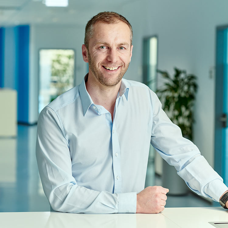 Lächelnder Mitarbeiter im hellblauen Hemd arbeitet an einem Laptop in einem modernen Büroflur.