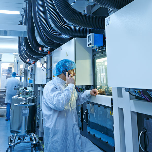 Techniker in Schutzkleidung und Haarnetz telefoniert und überwacht automatisierte Anlagen in einer Reinraum-Produktionsanlage.