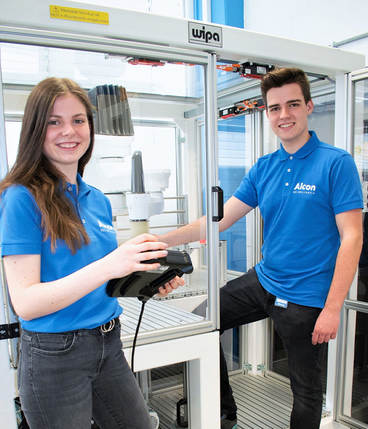 Zwei junge Menschen in blauen Alcon-Poloshirts stehen in einer modernen Trainings- oder Fabrikumgebung. Eine Person hält ein Steuergerät, während die andere selbstbewusst neben der Maschine steht. Beide lächeln in die Kamera.