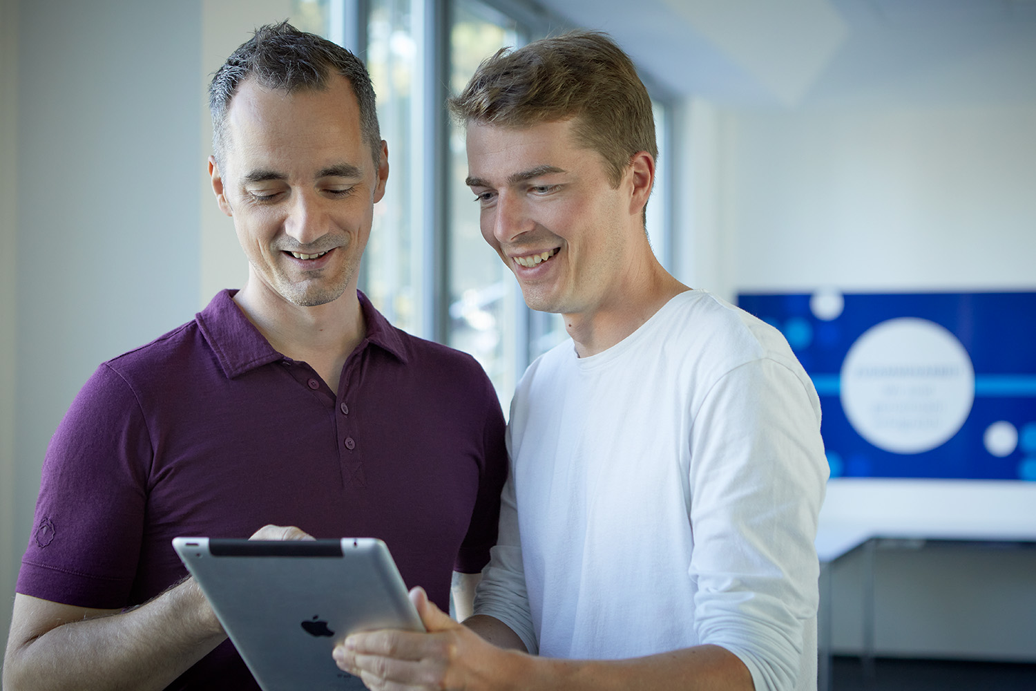Zwei Kollegen lächeln, während sie gemeinsam auf ein Tablet in einer modernen Büroumgebung schauen.
