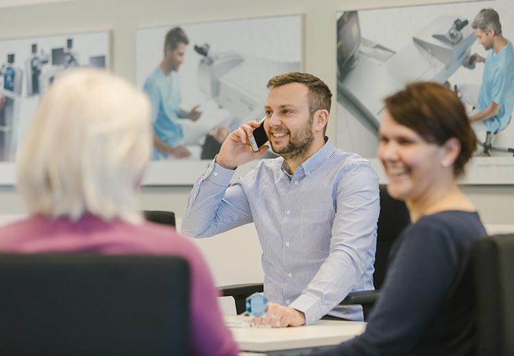 Mann im blauen Hemd lächelt, während er telefoniert, sitzt mit Kollegen in einer Büroumgebung.