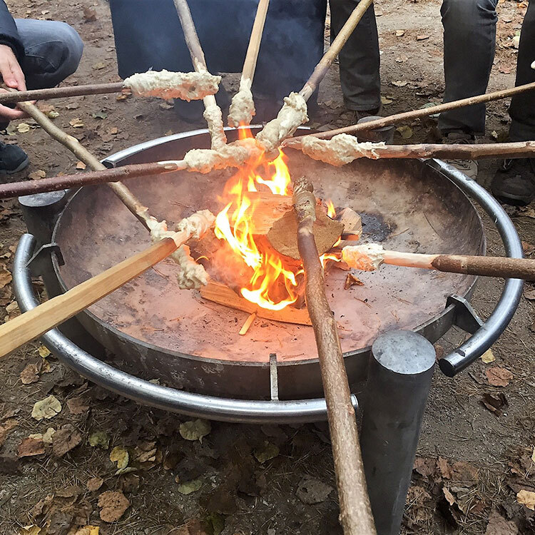 Gruppe von Menschen röstet Stockbrotteig über einem offenen Lagerfeuer in einer Metallfeuerschale im Freien.