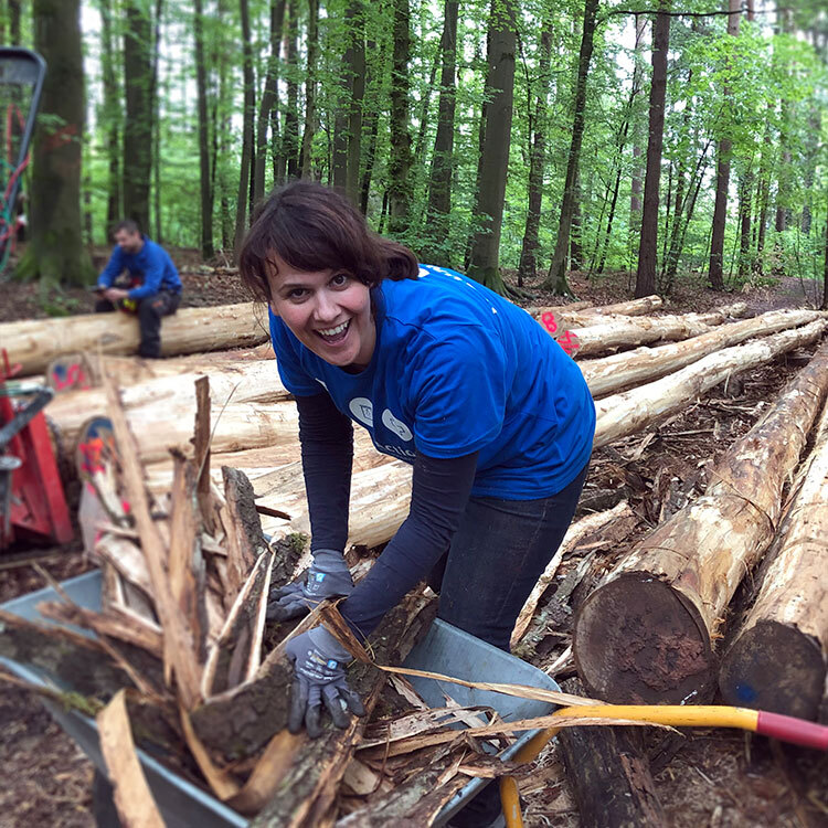 Person im blauen T-Shirt lächelt, während sie Holzstücke in eine Schubkarre im Wald sammelt, mit Baumstämmen und einer weiteren arbeitenden Person im Hintergrund.