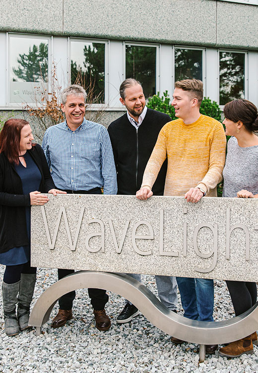 Gruppe von WaveLight-Mitarbeitenden, die zusammen vor dem Firmengebäude neben dem WaveLight-Schild stehen und lächeln.